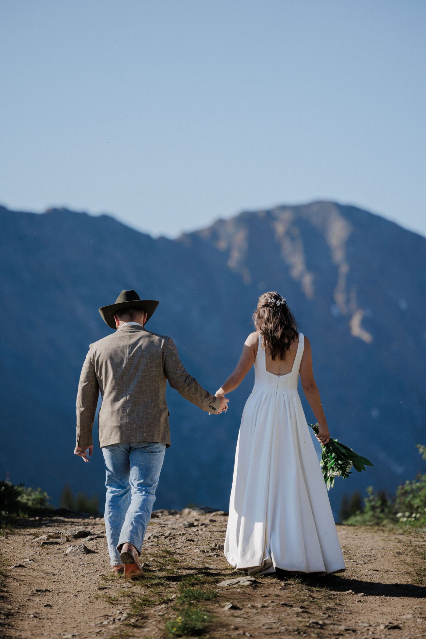 bride and groom hold hands and walk along trail during colorado summer wedding