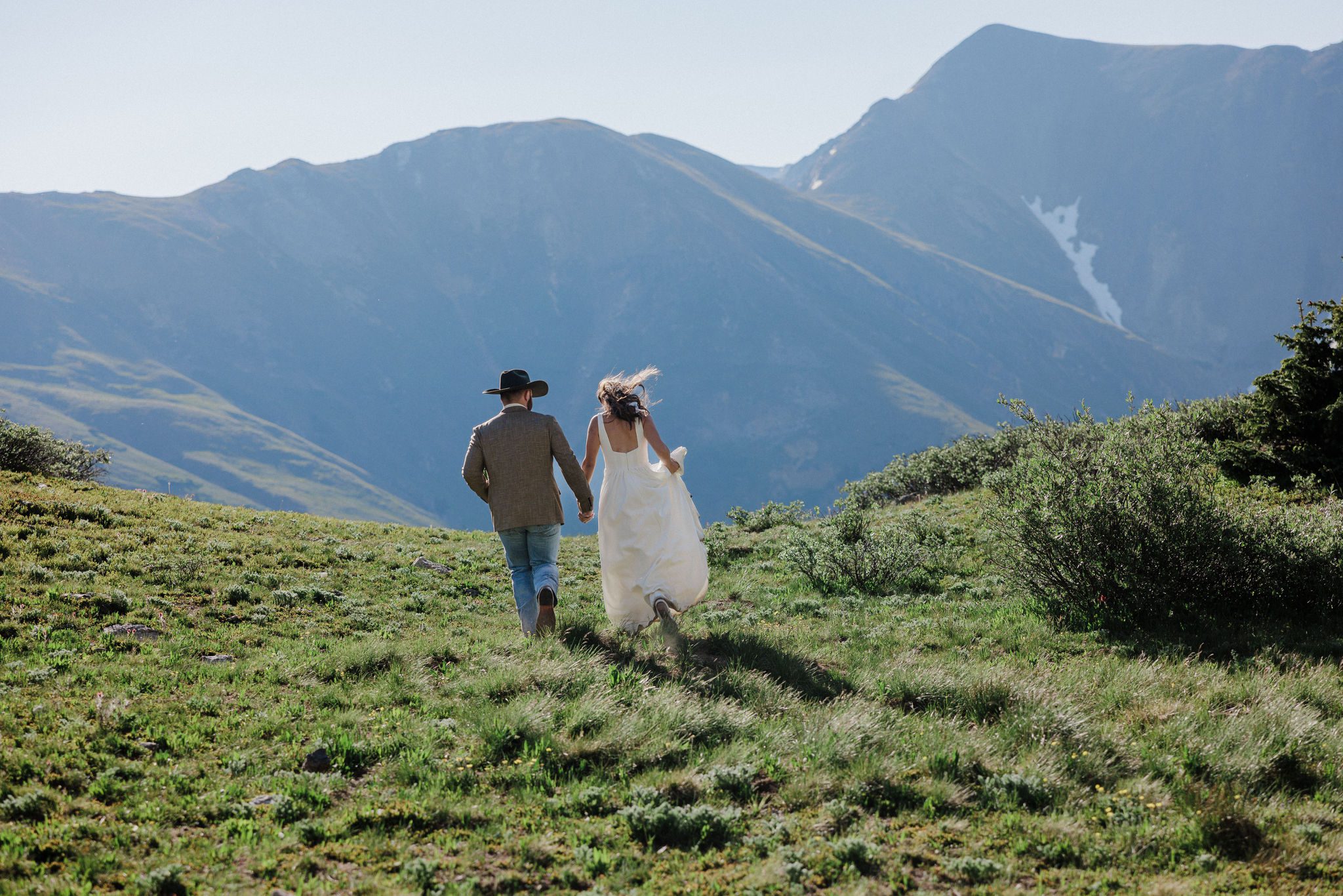 bride and groom run through field in the mountains during summer colorado wedding
