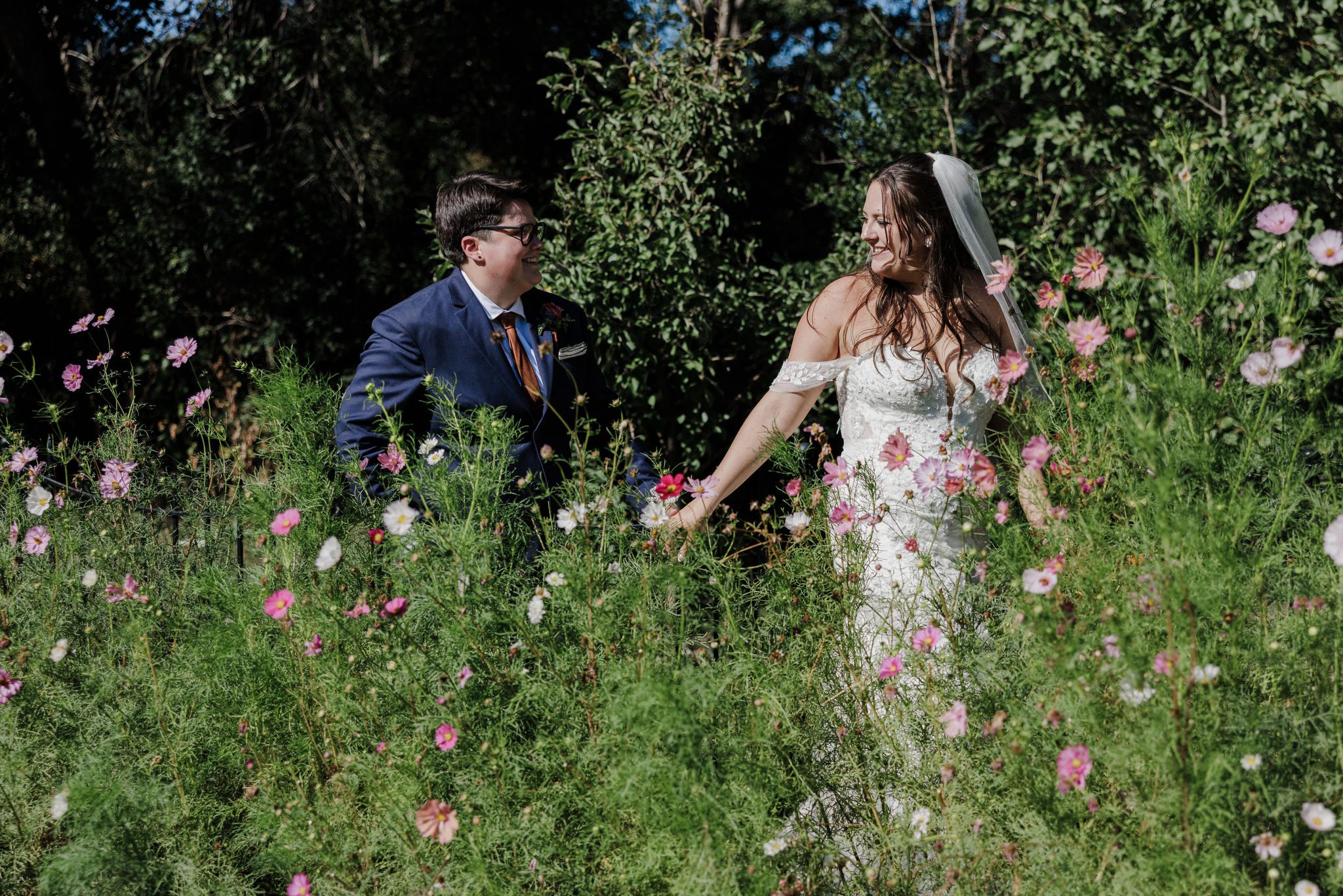 two brides hold hands and walk through wildflowers during summer wedding in colorado