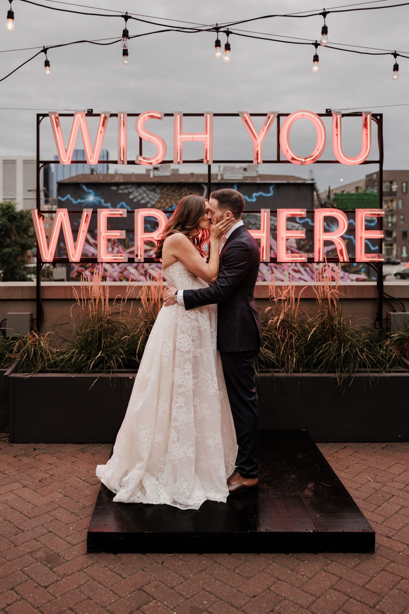 bride and groom kiss in front of a 'wish you were here' neon sign at summer wedding venue in colorado