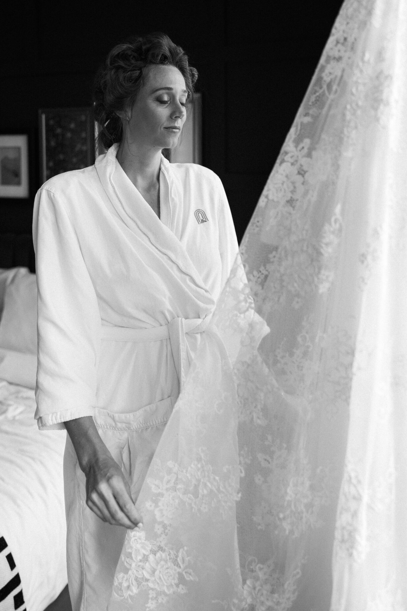 bride stands in robe and touches wedding dress that is hanging in hotel window