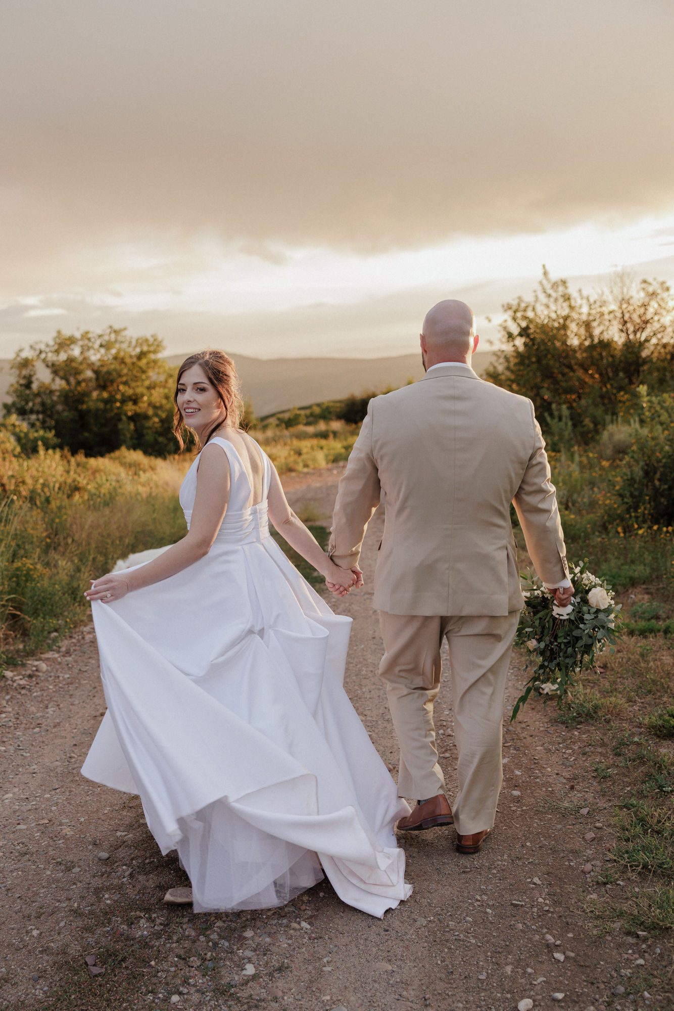 bride and groom hold hands and walk along trail at colorado airbnb during micro wedding reception in the summer