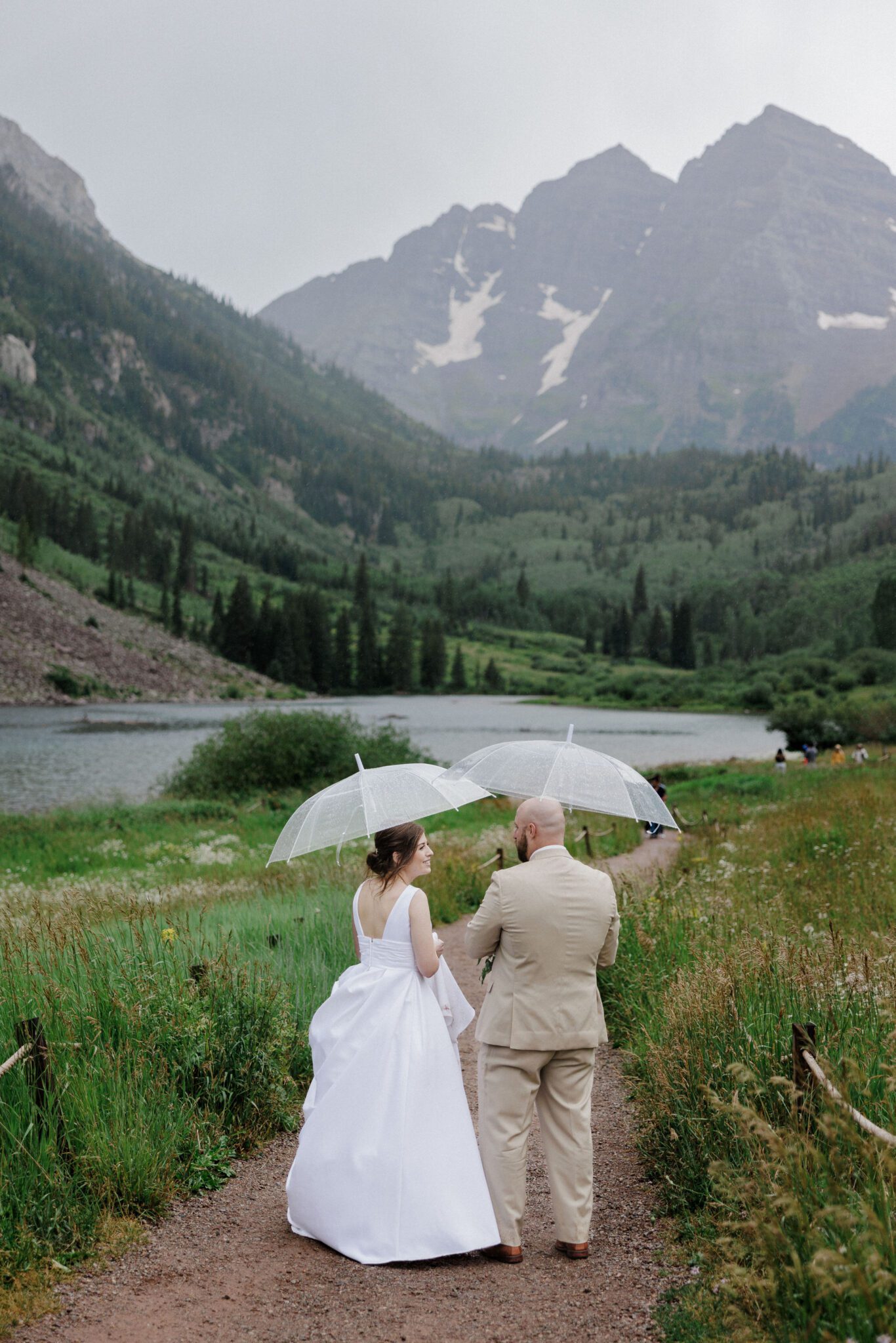 bride and groom hold umbrellas and walk on trail during colorado mountain lake wedding