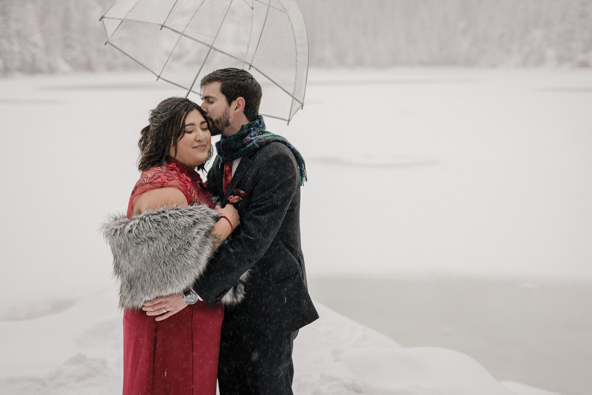 groom kisses brides head under clear umbrella during their winter mountain lake elopement at RMNP