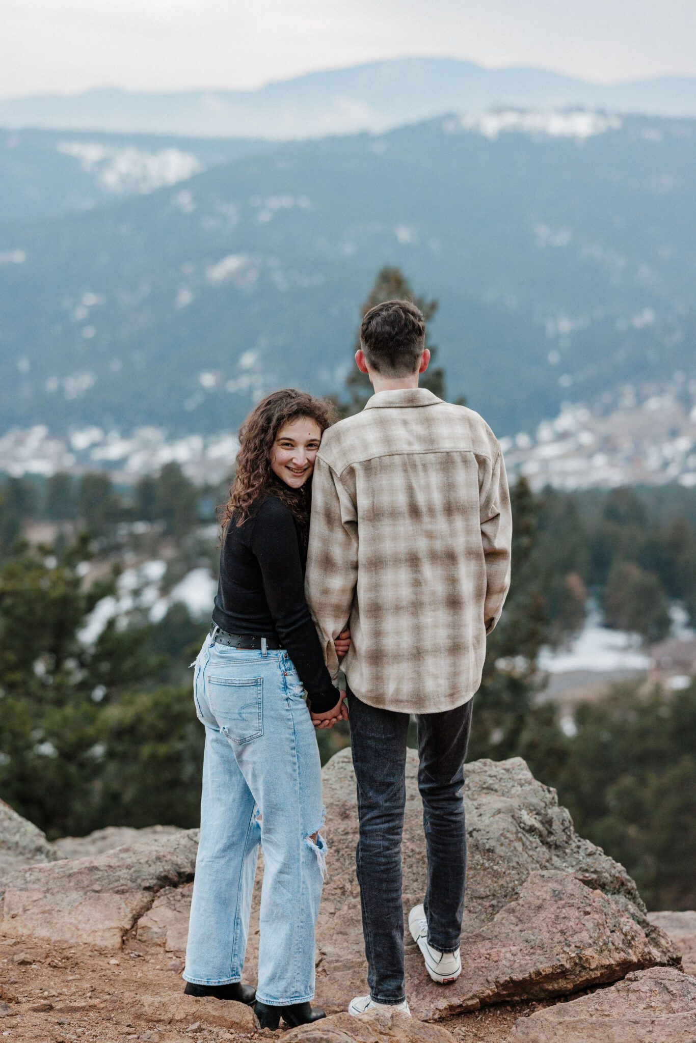 man and woman stand on the edge of a rock at mt falcon and pose during colorado engagement photos 