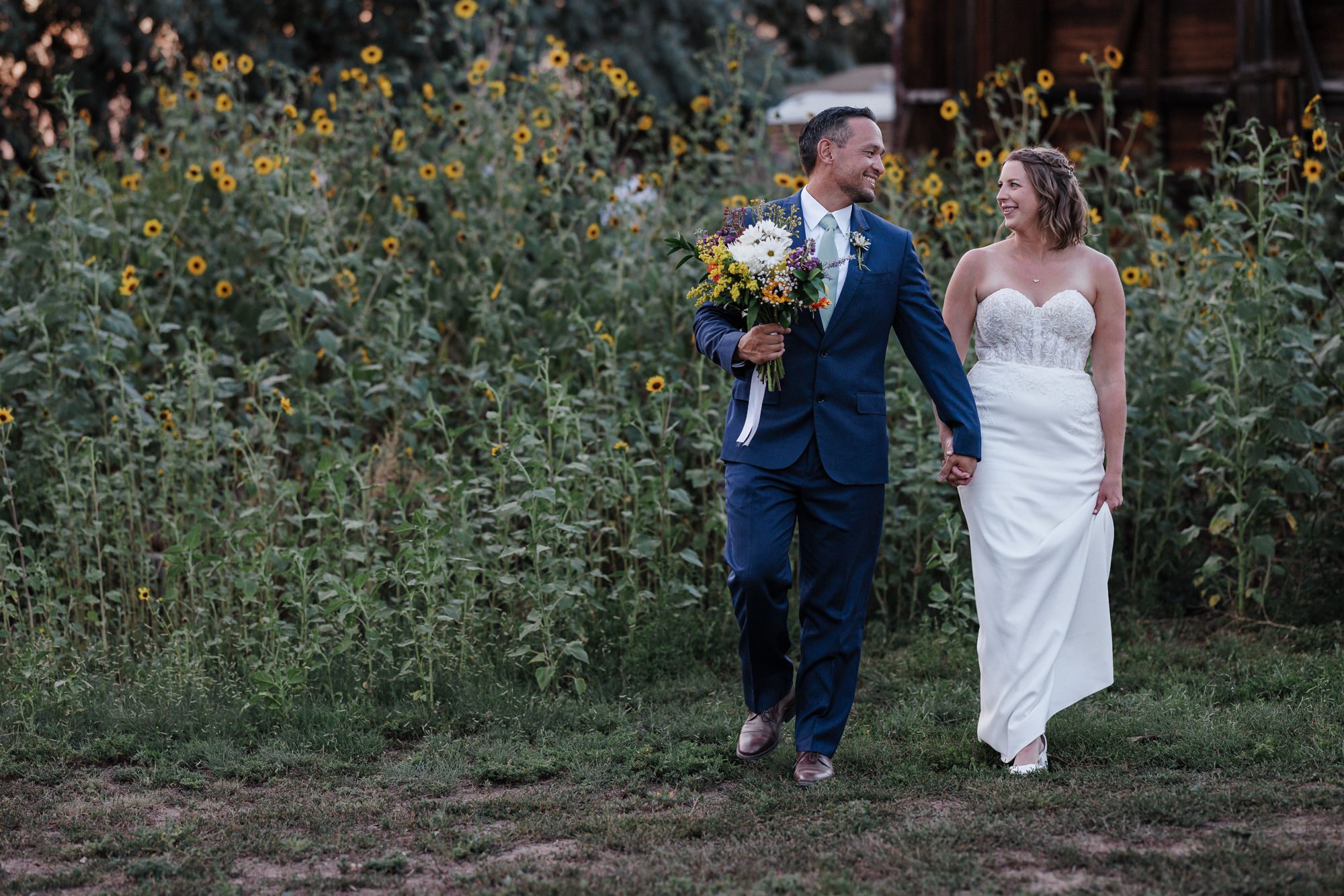 bride and groom hold hands and walk through sunflower field