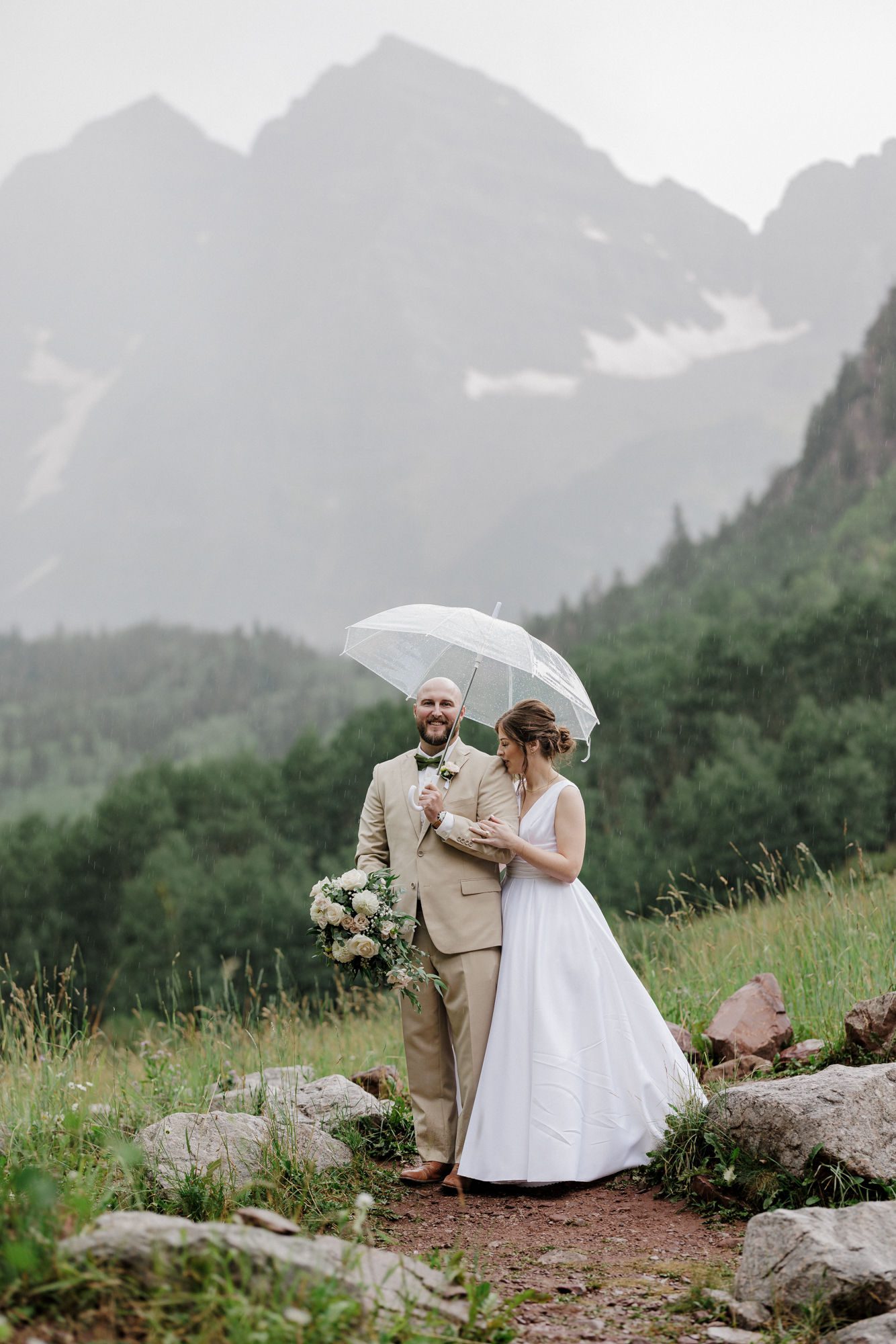 bride and groom stand at Maroon Bells in the rain, with clear umbrellas brought by micro wedding planner.