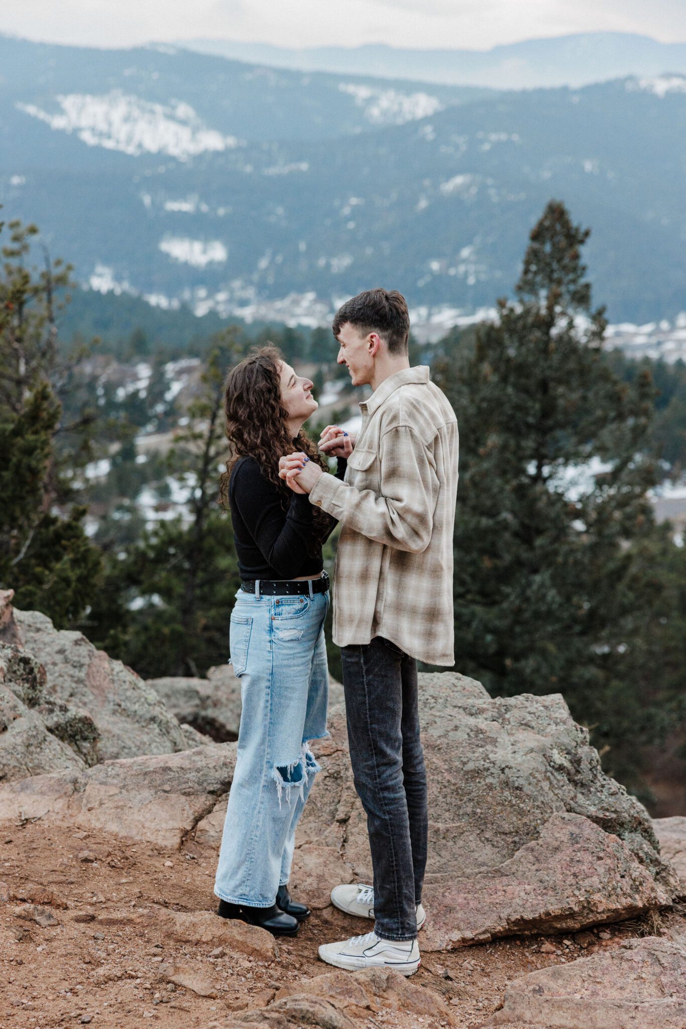 engaged couple smiles at each other and grabs hands after proposal in the mountains