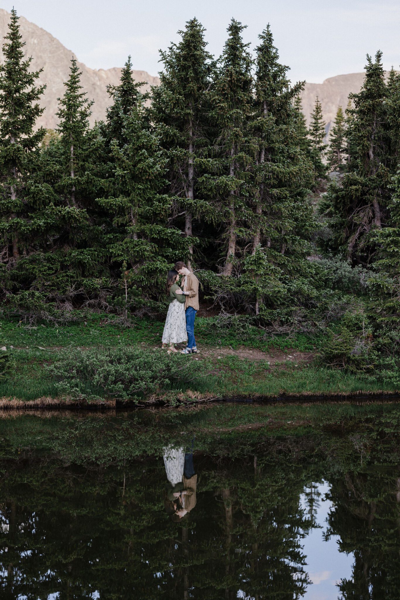 engaged couple embraces beside an alpine lake and evergreens during proposal photos