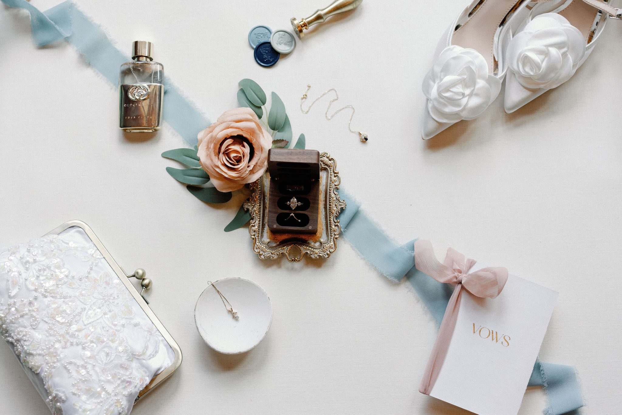 wedding detail flat lay photo with ring in ring box, wedding heels, vow book, white clutch, and perfume