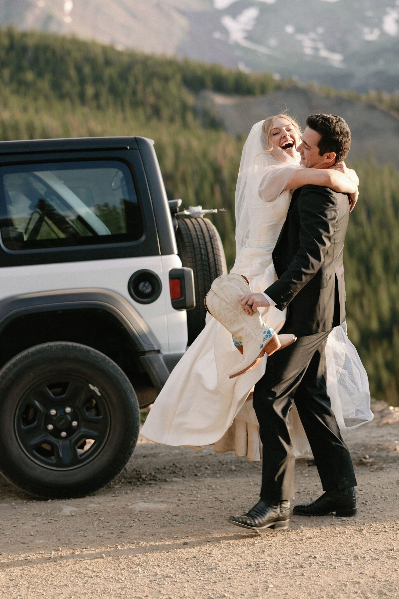 groom holds up bride and her shoes and walks to their jeep