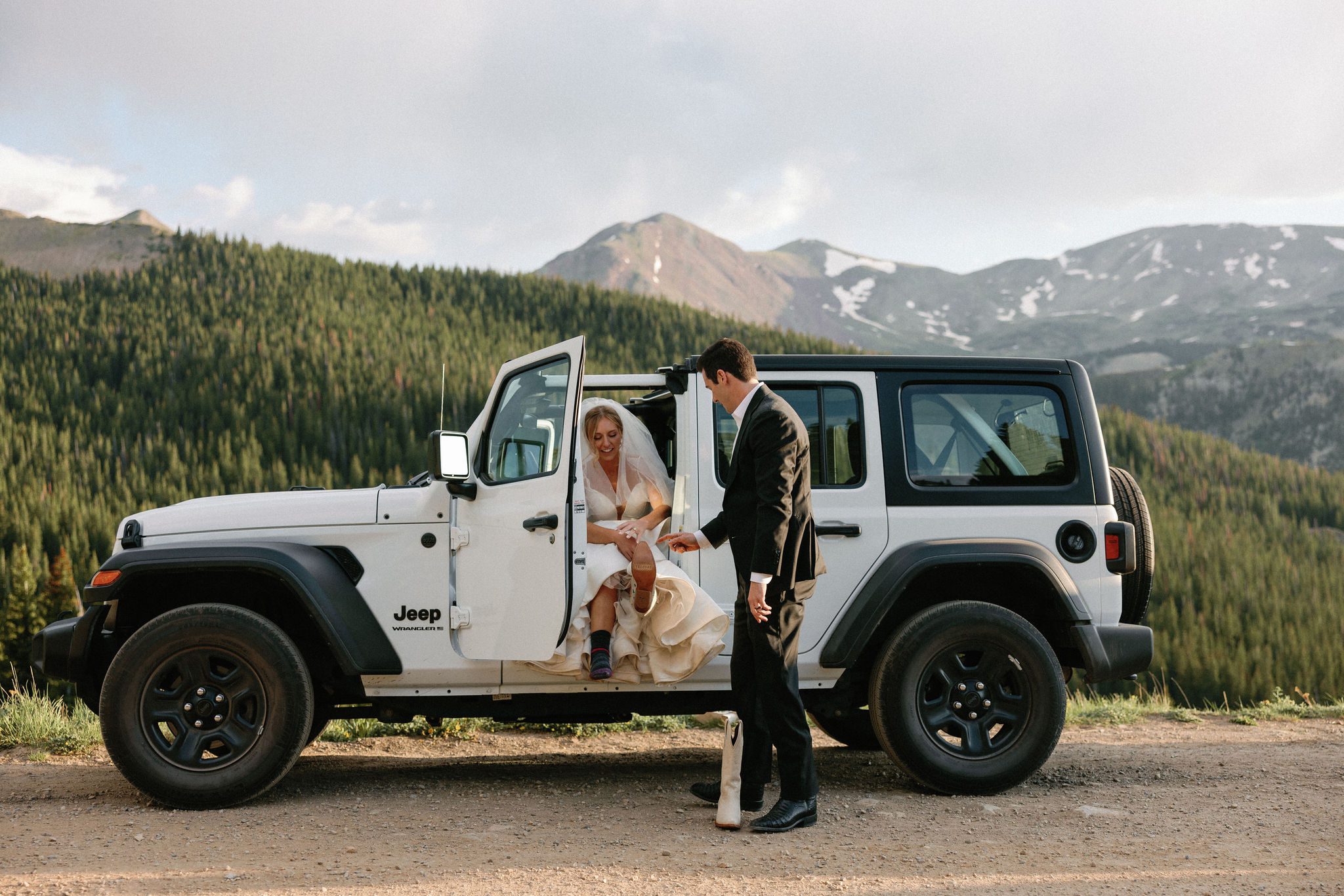 bride sits in their white jeep as groom helps her put her white boots on