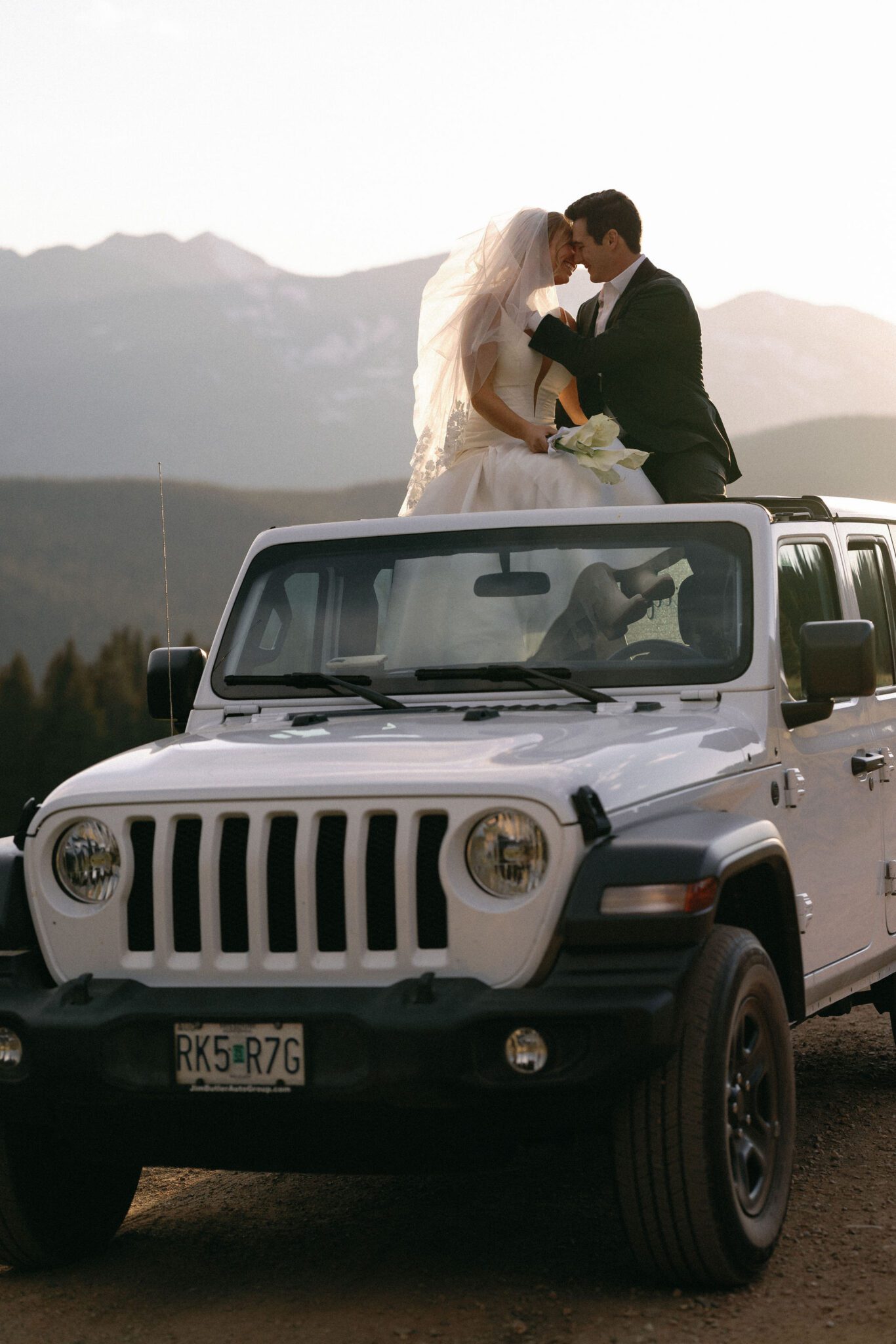 bride and groom sit on the sunroof of a white jeep and take sunset photos after their airbnb wedding in colorado