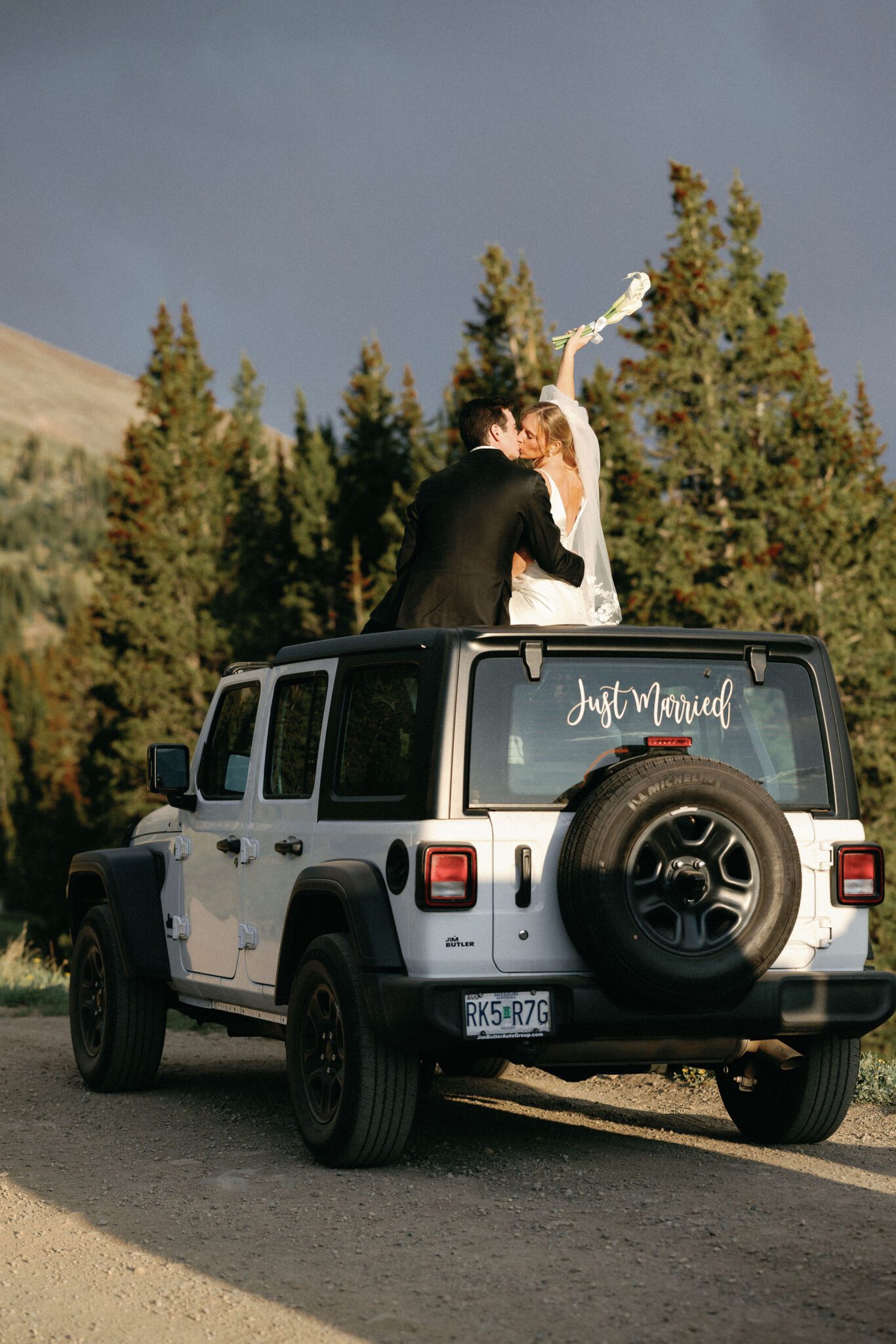 bride and groom sit on the sunroof of a white jeep, her hand is in the air, with 'just married' decal on the back window