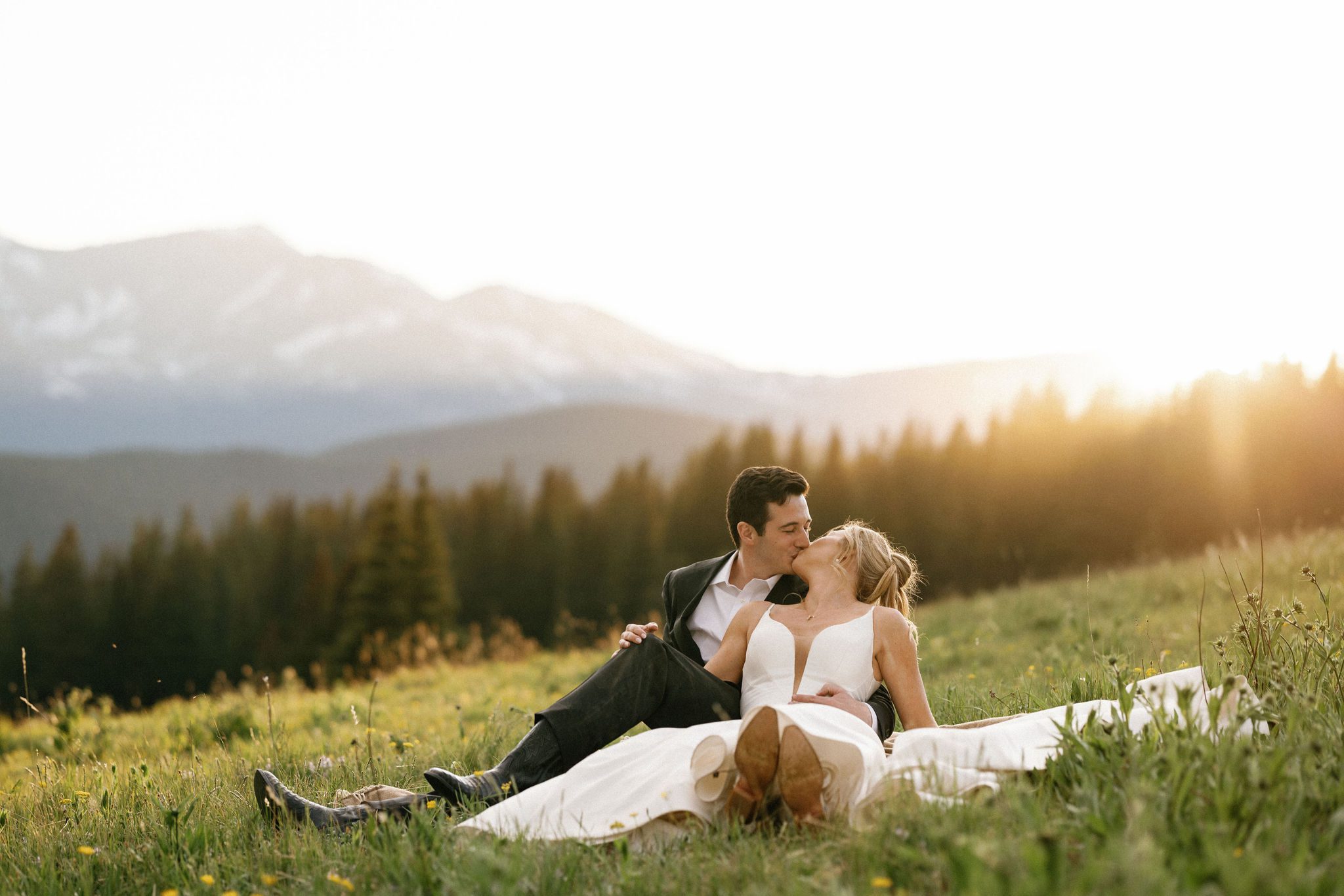 during wedding photos, bride and groom sit in the grass and share a kiss during sunset