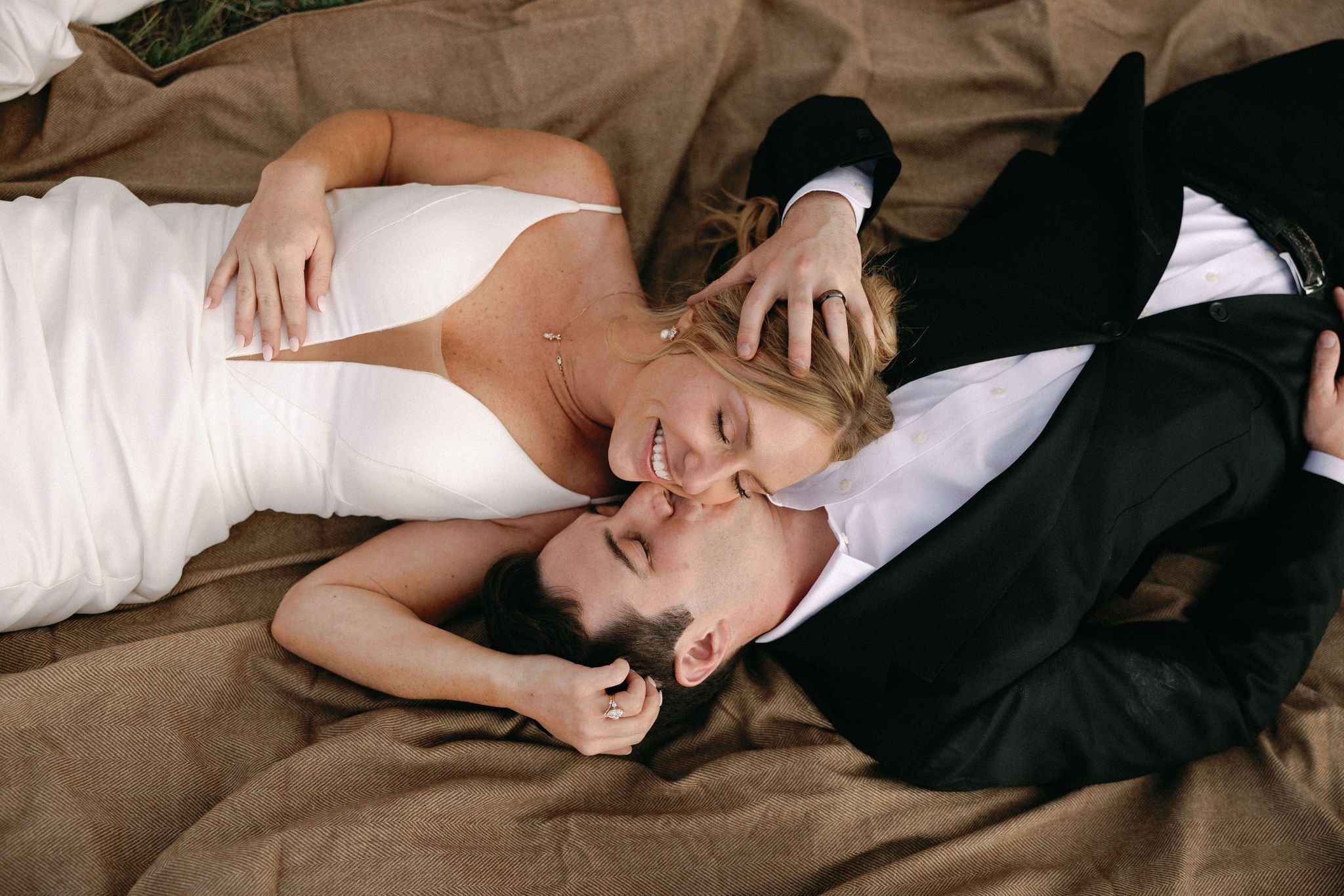 bride and groom lay on their backs on a blanket and groom reaches over to kiss the brides cheek