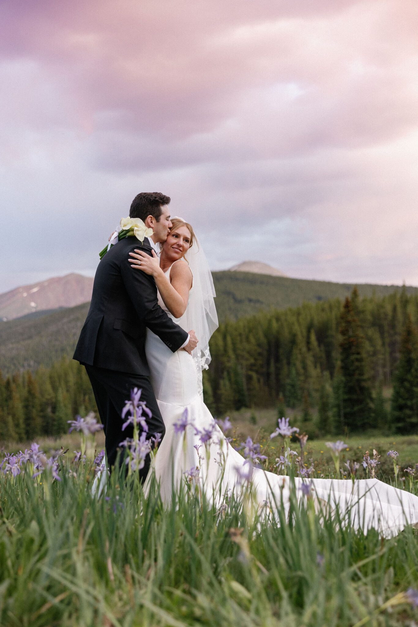 after their colorado airbnb wedding, bride and groom stand in the mountain wildflowers as groom kisses the brides head