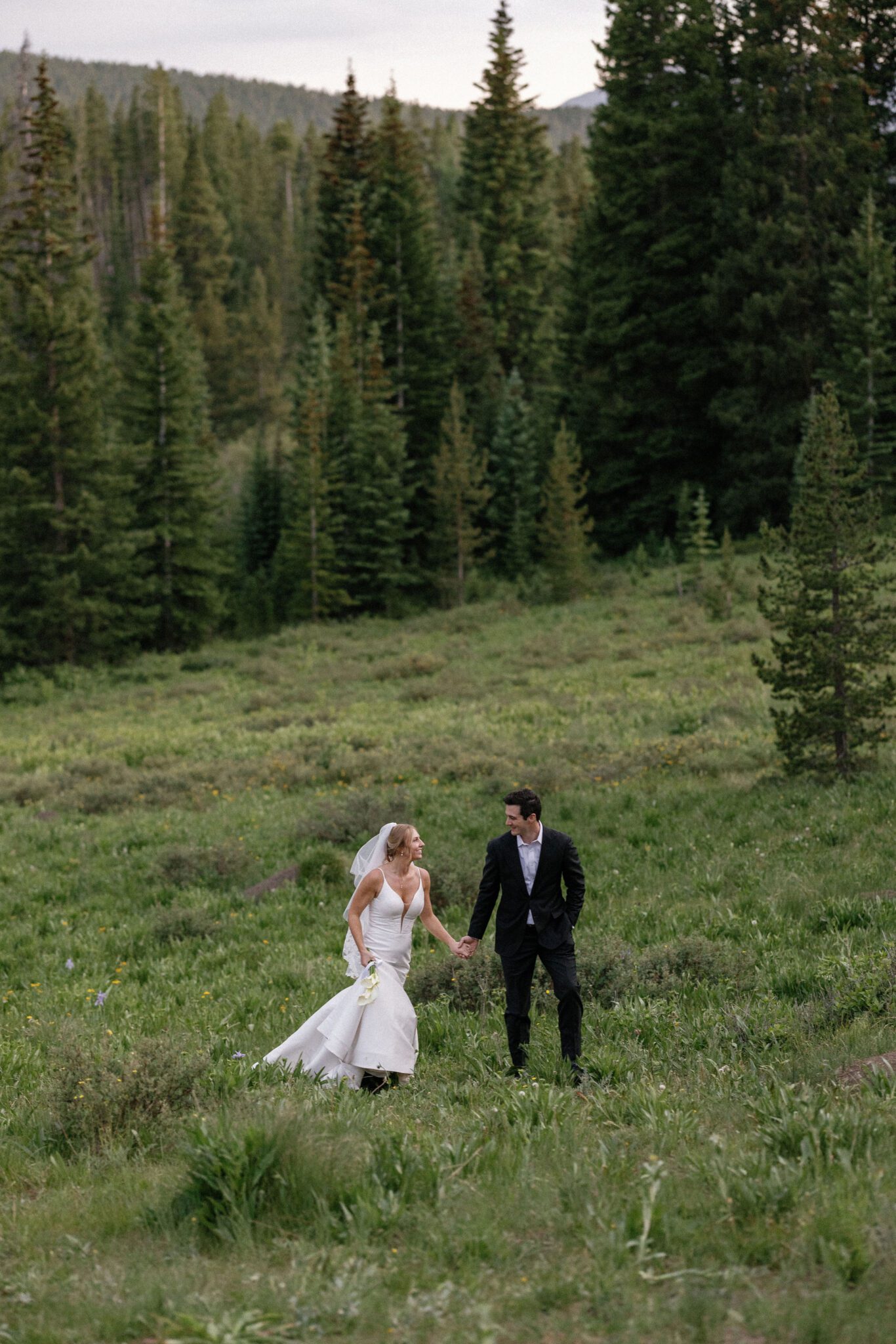 bride and groom hold hands and walk through a field in the mountains of colorado after airbnb wedding