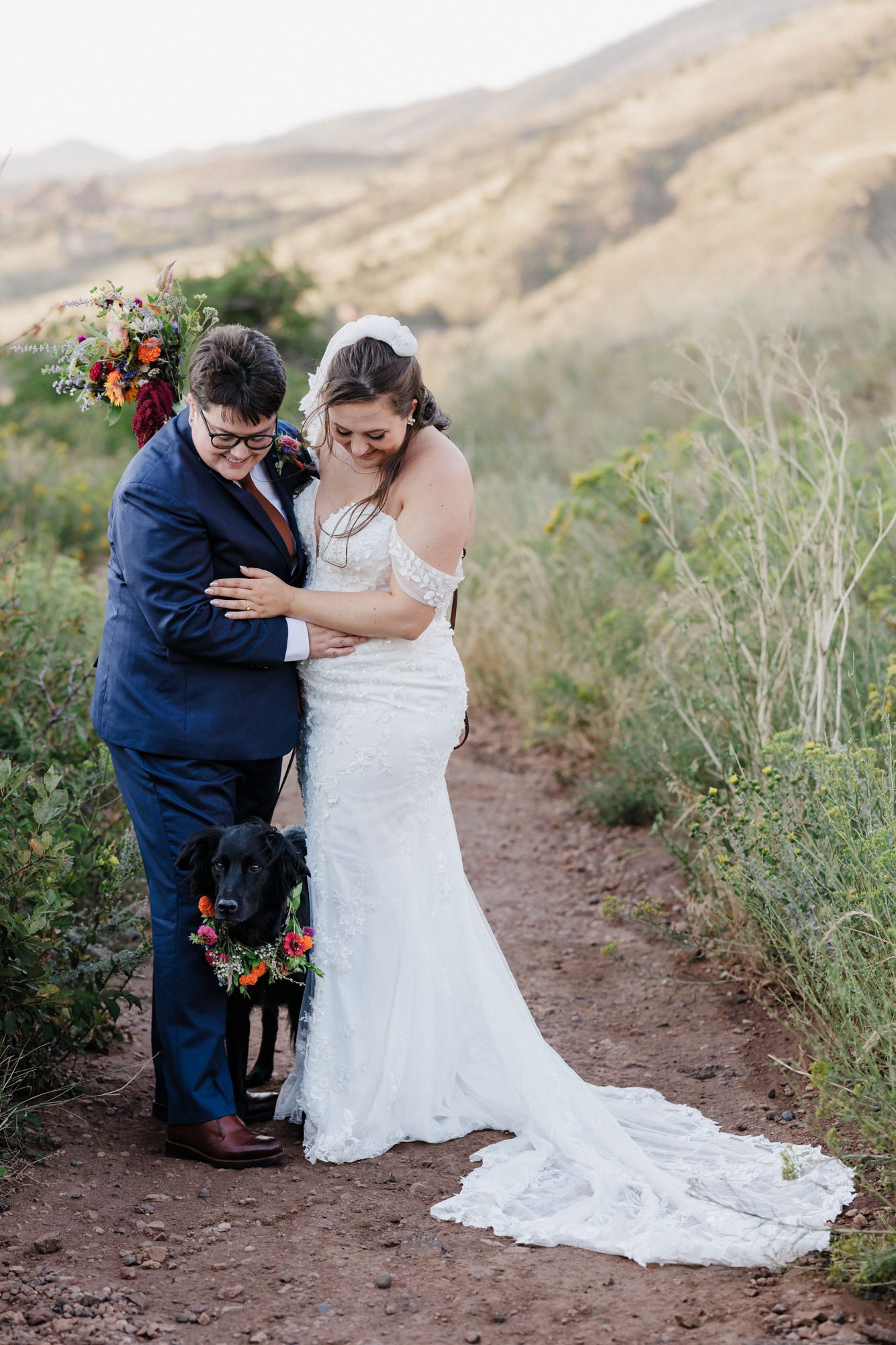two brides hug while they look down at their dog. they are standing on a trail at red rocks park