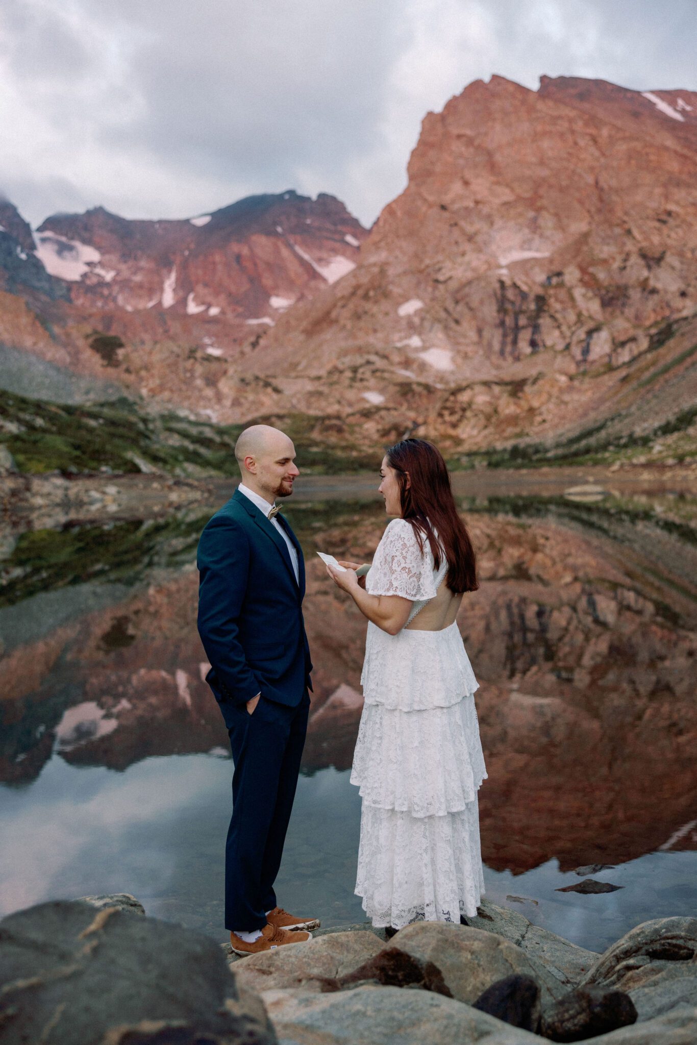 bride and groom stand in front of alpine lake during sunrise at their colorado elopement location, lake isabelle